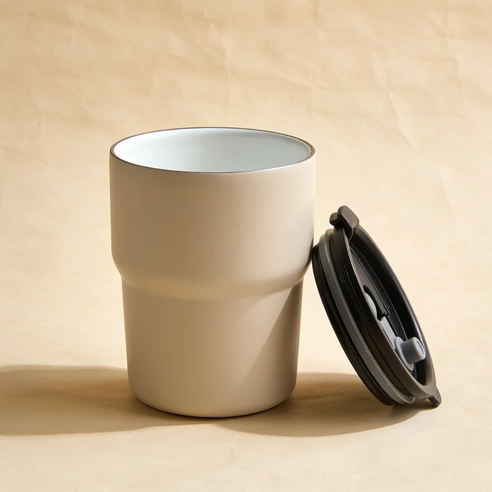 Short sand beige stainless steel travel cup without handle, clearly visible white ceramic-lined interior at rim, matching black removable drinking lid placed next to cup